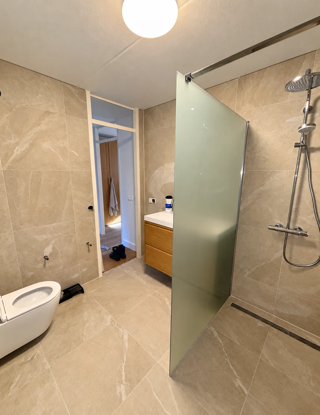 Badkamer met beige tegels, glazen douchewand, douche en wastafelmeubel